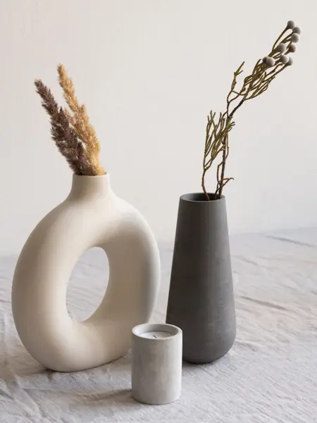 Two ceramic handmade vases with dried wildflowers and spikes and aromatic candle standing on table covered with white linen cloth