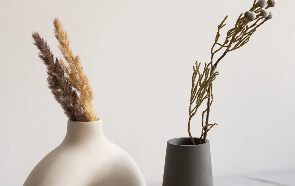 Two ceramic handmade vases with dried wildflowers and spikes and aromatic candle standing on table covered with white linen cloth