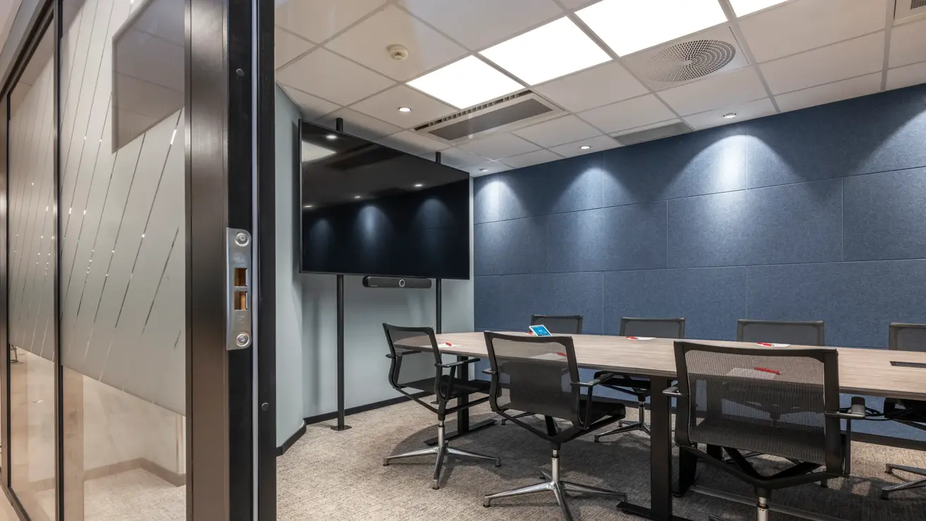 Møterom med styrebords oppsett sett fra inngangsdøren. Blå vegg og stor skjerm på veggen.