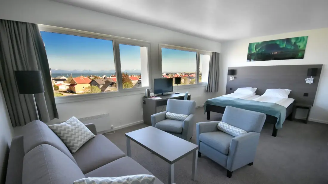 Business-rom med sofa, salongbord, to hvilestoler, dobbeltseng, TV, panoramautsikt mot bebyggelse og sjø.