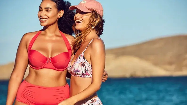 To smilende damer kledd i bikini med sjø og fjell i bakgrunnen