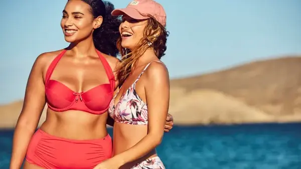 To smilende damer kledd i bikini med sjø og fjell i bakgrunnen