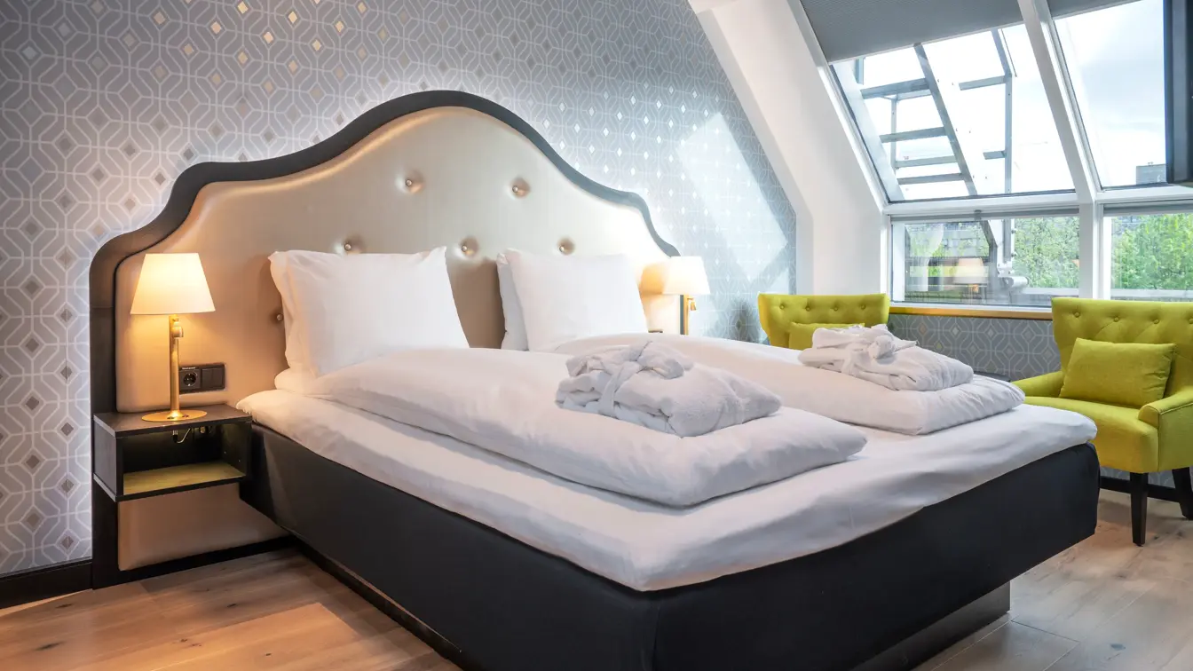 Dobbeltseng med morgenkåper, mønstret sølv tapet, gule stoler og store vinduer i soverom i junior suite på Thon Hotel Cecil