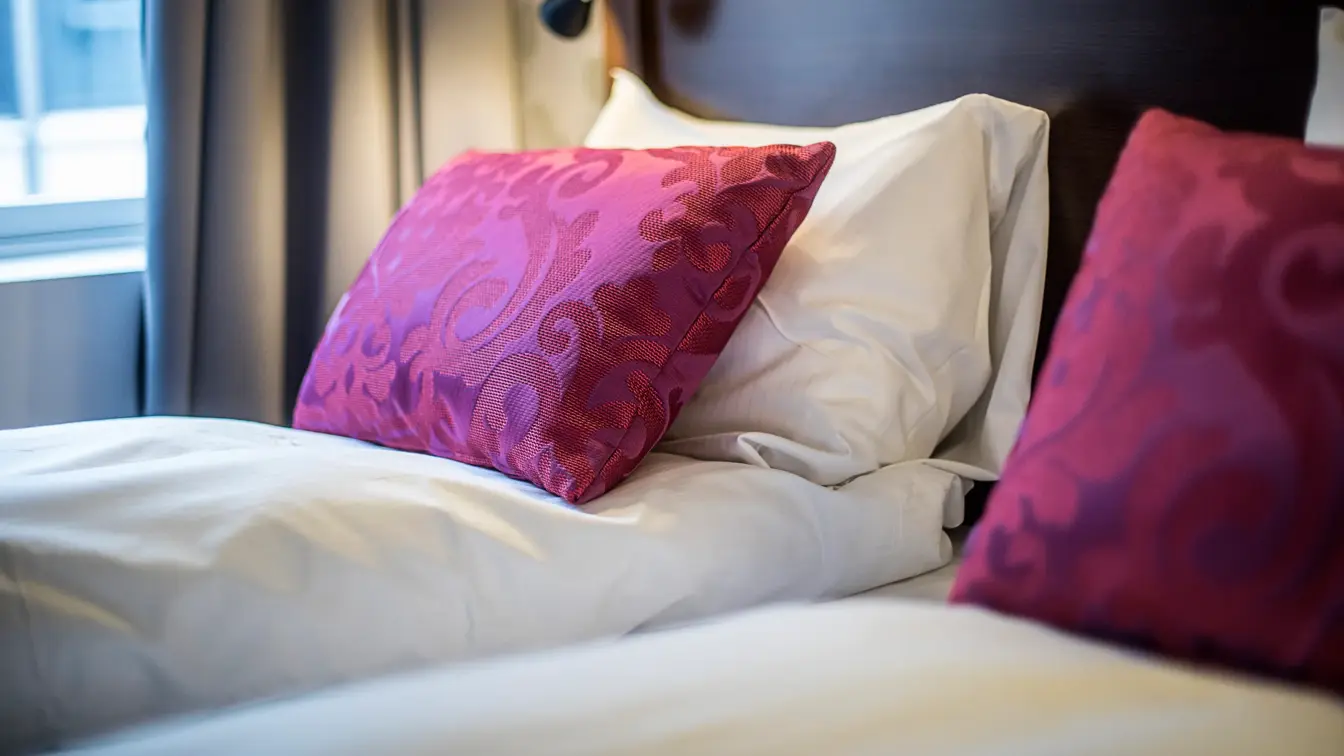 Dobbeltseng, rosa pynteputer, vindu, grå gardin. 