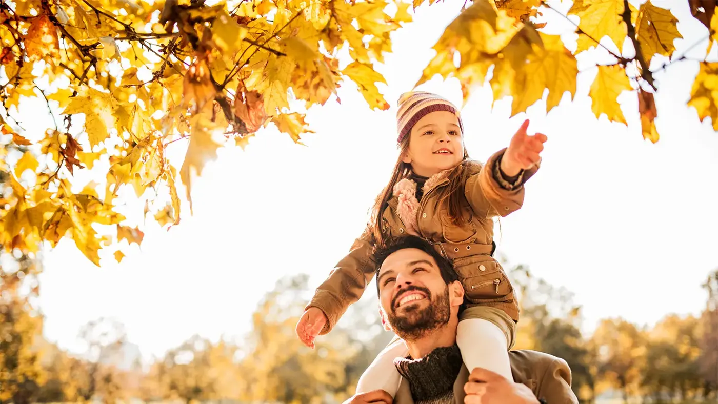 jente sitter på skuldrene til faren sin og strekker seg etter høstløvet som henger ned fra et tre