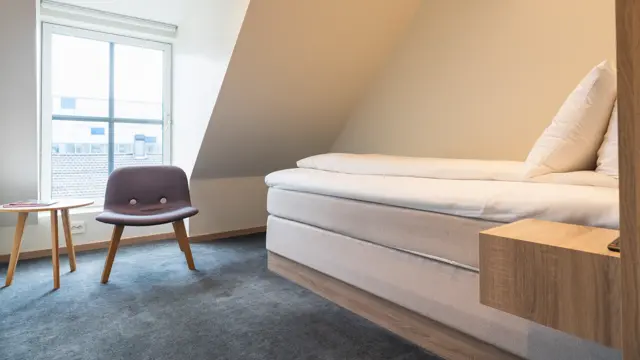 Enkeltrom med enkeltseng, teppegulv, lenestol med stort vindu bak.