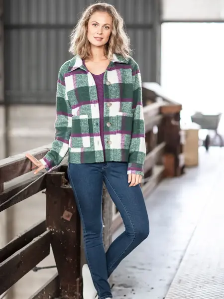 Dame med lyst hår står lent mot et gjerde. Har på seg blåe jeans, lilla genser med en grønn, hvit og lilla-rutete skjortejakke