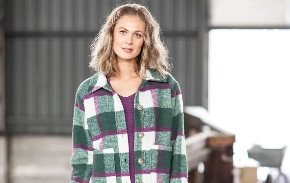 Dame med lyst hår står lent mot et gjerde. Har på seg blåe jeans, lilla genser med en grønn, hvit og lilla-rutete skjortejakke