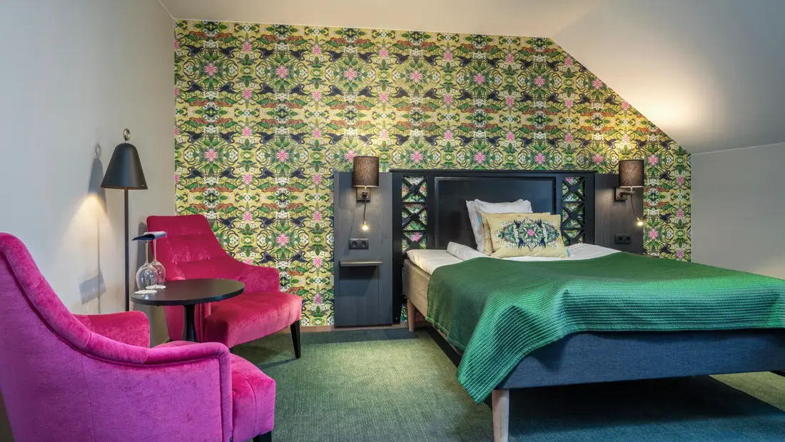 Superorrom med dobbeltseng, to, rosa hvilestoler, mønstrete fargerik tapet og vegg-til-vegg-teppe.