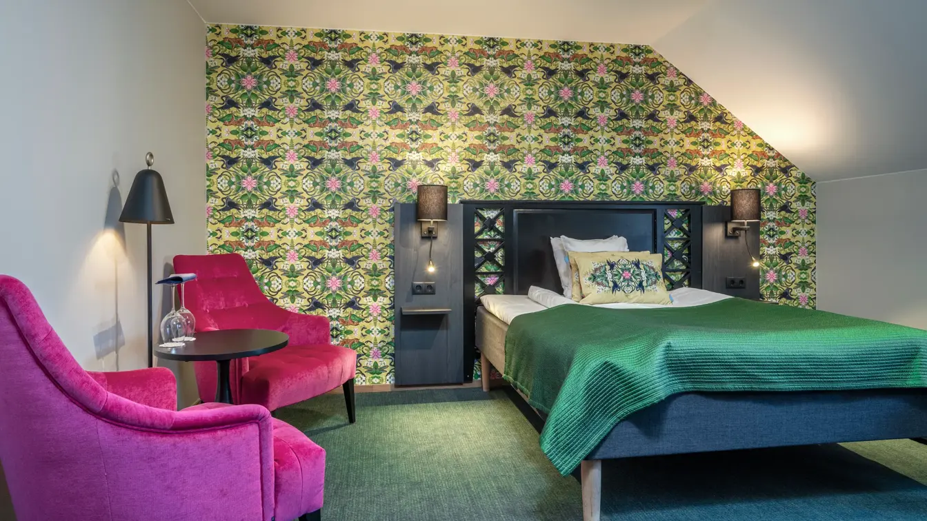 Superorrom med dobbeltseng, to, rosa hvilestoler, mønstrete fargerik tapet og vegg-til-vegg-teppe.