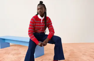 En kvinne i rød genser med striper og blå jeans sitter på en blå benk.