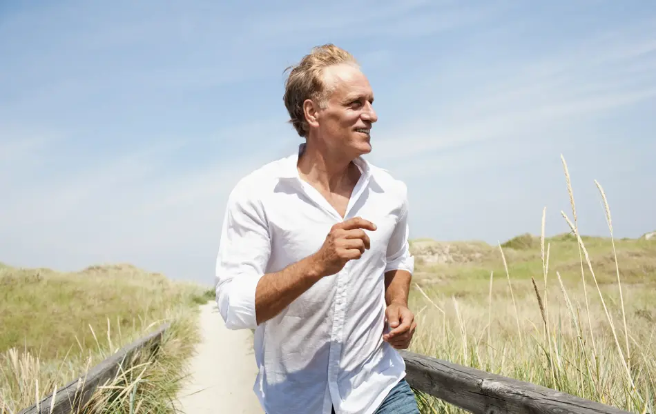Eldre mann på strand iført hvit skjorte. Sand og gress og blå himmel.