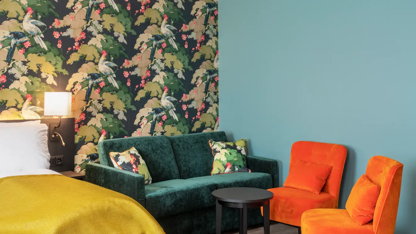 Seng med gult sengeteppe. Fargerik tapet med fugler og blomster, grønn sofa, oransje stoler og mørkt bord i business rom på Thon Hotel Bristol Bergen
