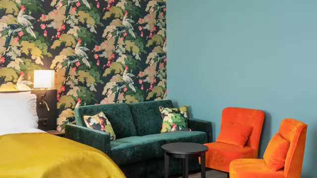 Seng med gult sengeteppe. Fargerik tapet med fugler og blomster, grønn sofa, oransje stoler og mørkt bord i business rom på Thon Hotel Bristol Bergen