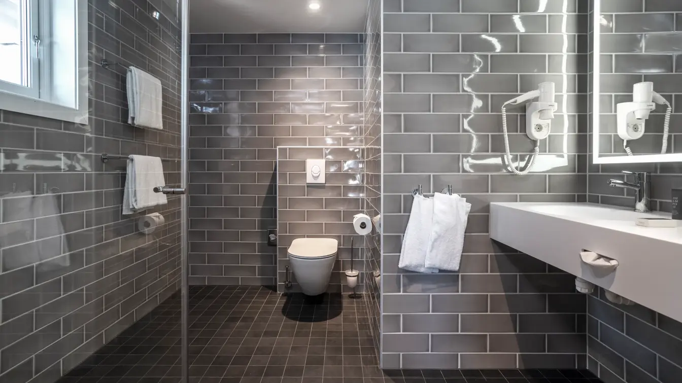 Grå veggfliser, svarte gulvfliser, vask, toalett, dusj, speil