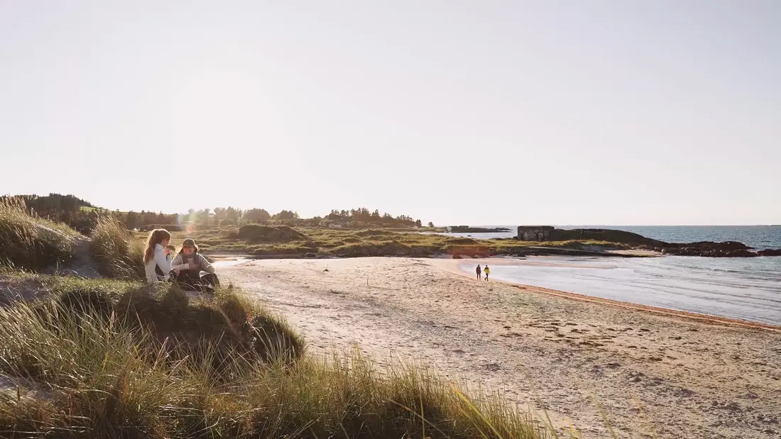 To kvinner sitter på vollen ved ei strand.