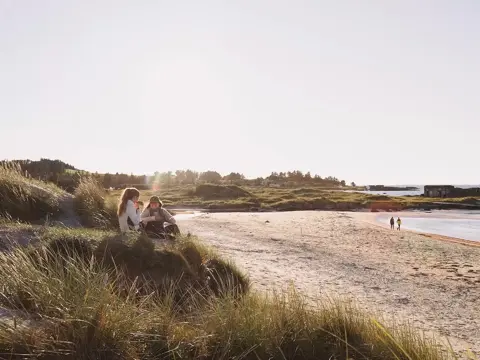 To kvinner sitter på vollen ved ei strand.