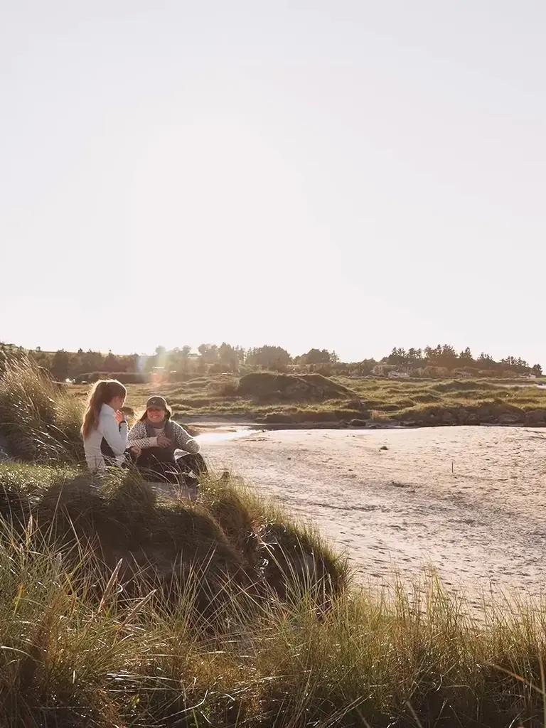 To kvinner sitter på vollen ved ei strand.