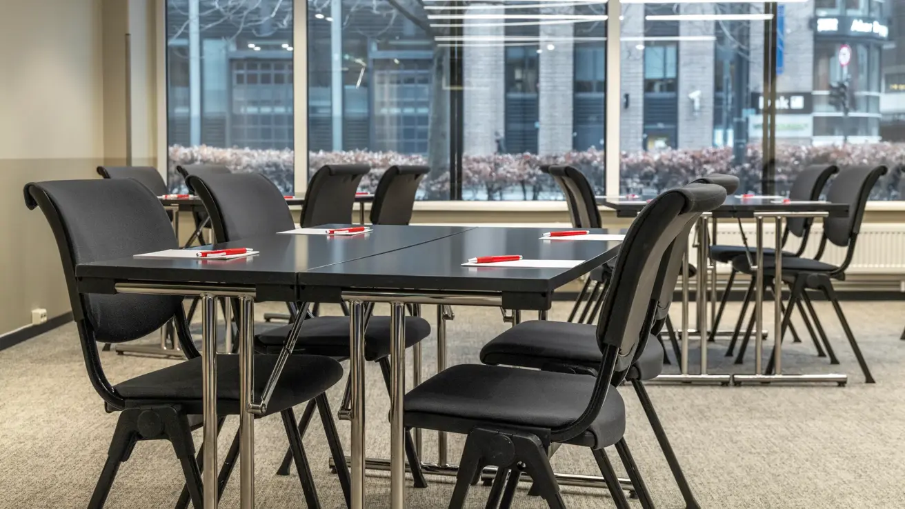 Nærbilde av møterom med flere gruppebord. Sorte stoler og bord, store vinduer langs en hel vegg ut mot gaten.