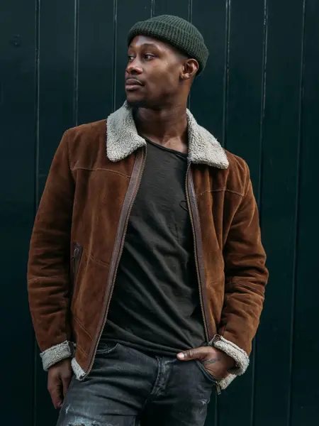 Mann i brun, semsket skinnjakke og grønn lue poserer foran vegg 