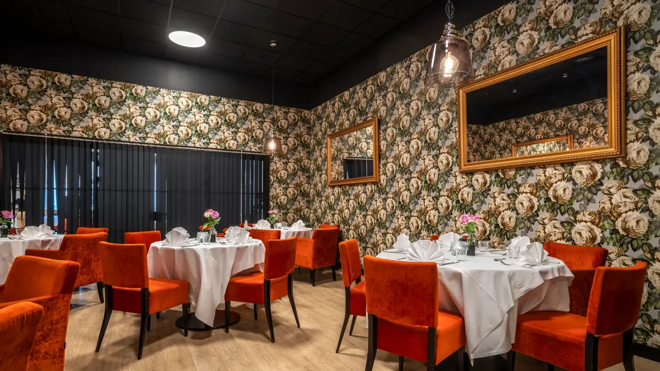 Chambre Separee i restaurant, med runde bord, hvite duker, oransje stoler, mønstrete tapet med avlange speil på veggen.