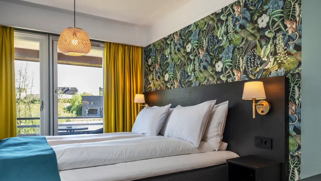 Standard rom med queen seng, blått sengeteppe, mønstrete og fargerik tapet, svart sengegavl, store vinduer, gule gardiner og balkong