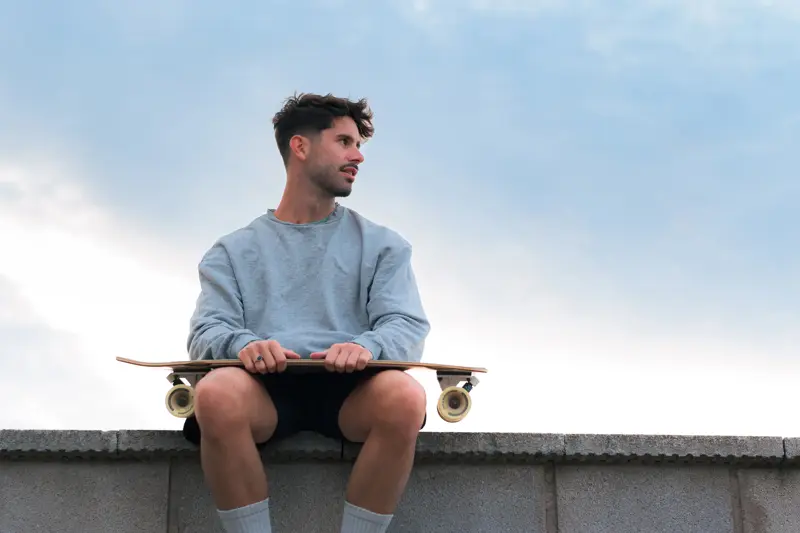 Mann med grå genser og skateboard sitter på murkant 