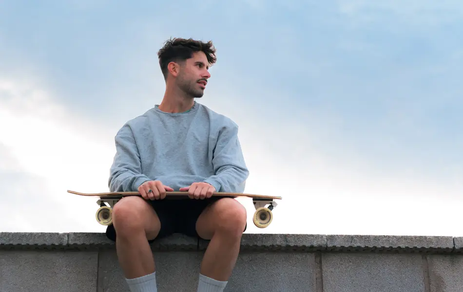 Mann med grå genser og skateboard sitter på murkant 