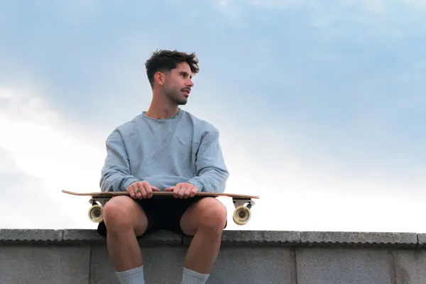Mann med grå genser og skateboard sitter på murkant 