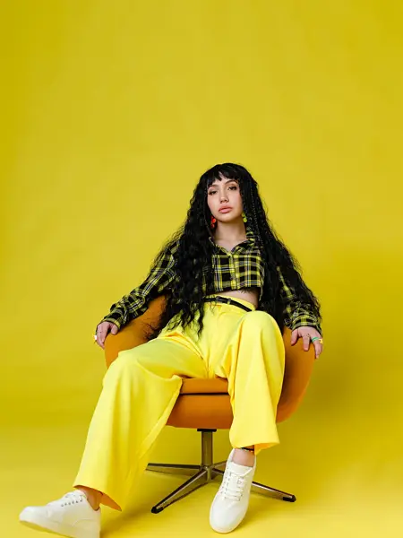 Jente med hvite sko, knallgul bukse og skjorte med gule og brune ruter sitter i en oransje stol og ser mot kamera, i et helt gult rom.