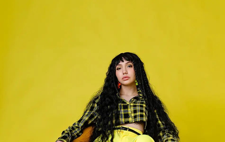 Jente med hvite sko, knallgul bukse og skjorte med gule og brune ruter sitter i en oransje stol og ser mot kamera, i et helt gult rom.