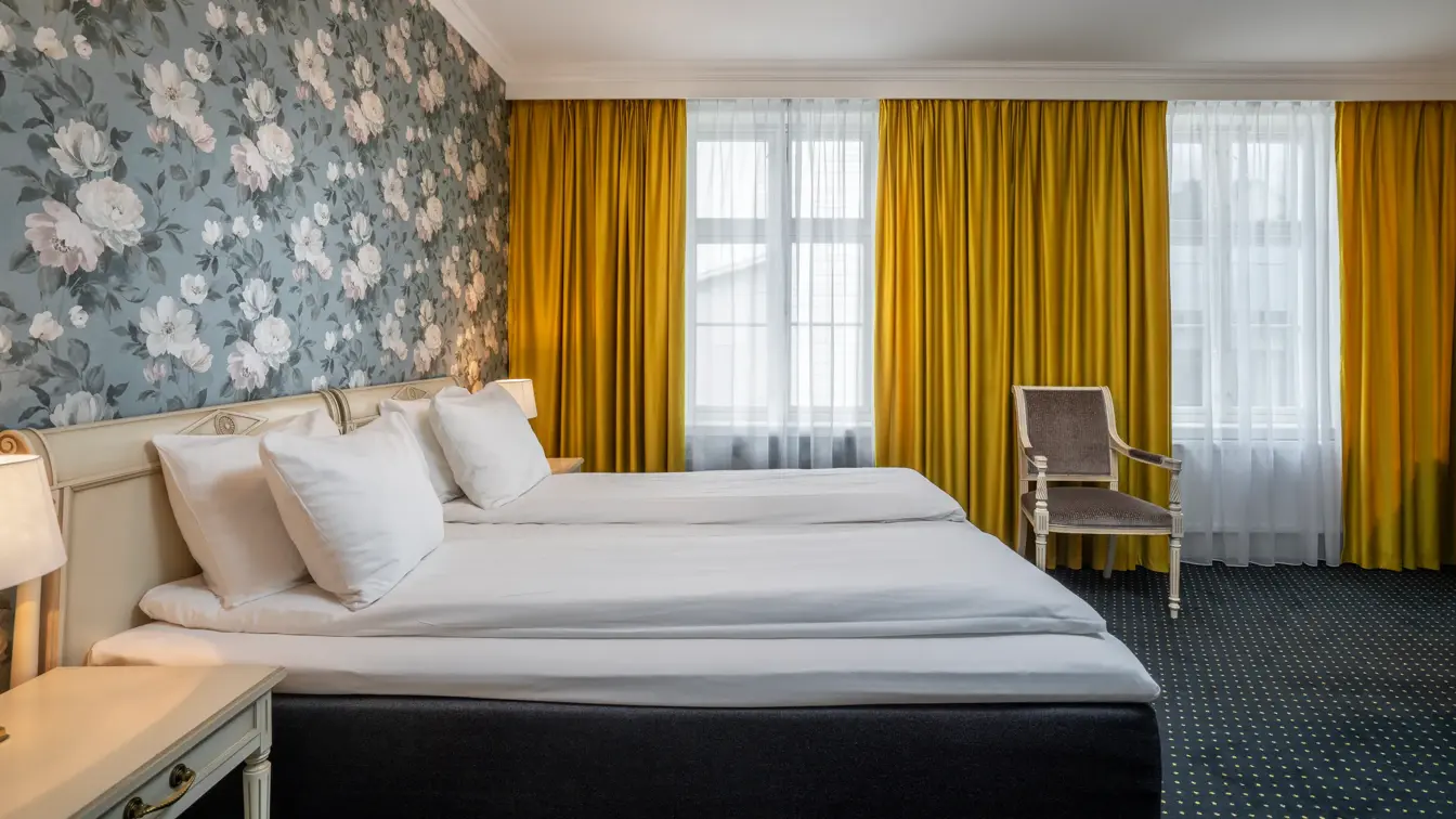 To enkeltsenger, nattbord, vindu, gule gardiner, tapetvegg, stol. 