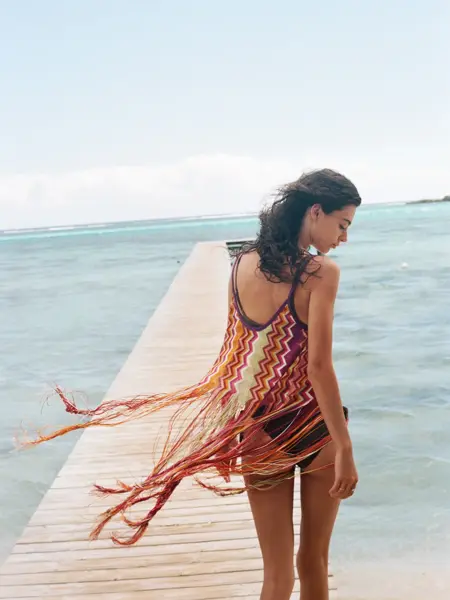 A young brunette girl walks along a beach pier on a sunny afternoon; her swimsuit coverup blows in the wind and she looks down contently at the ocean water