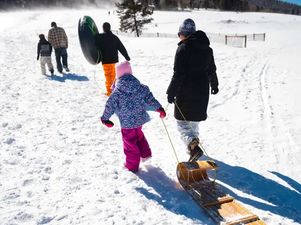 Voksne og barn sett bakfra mens de går i snøen og drar på kjelke og bærer badering til å ake på