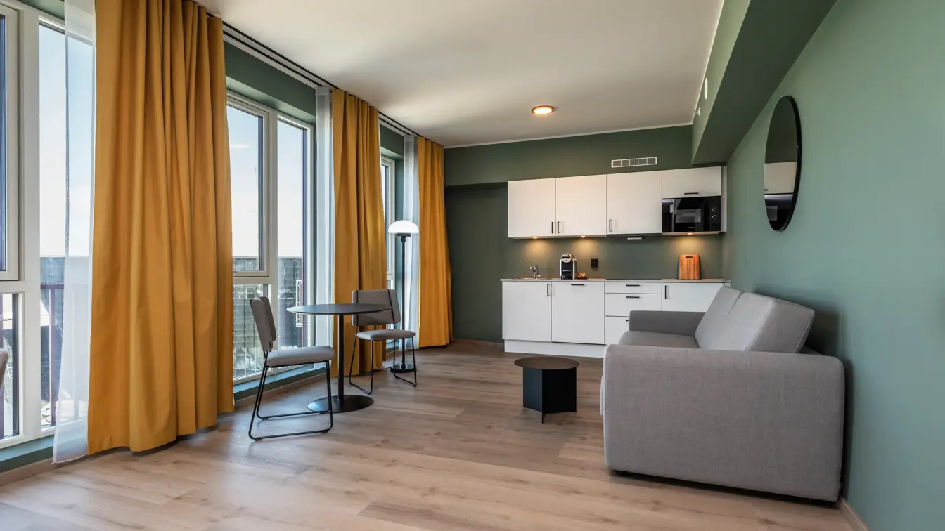 Business rom med kjøkken krok, grå sofa, spiseplass, grønne vegger, gule gardiner og parkett.