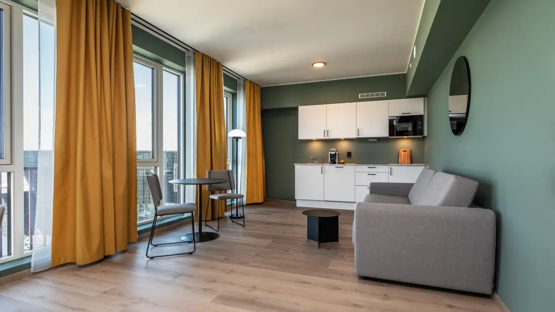 Business rom med kjøkken krok, grå sofa, spiseplass, grønne vegger, gule gardiner og parkett.