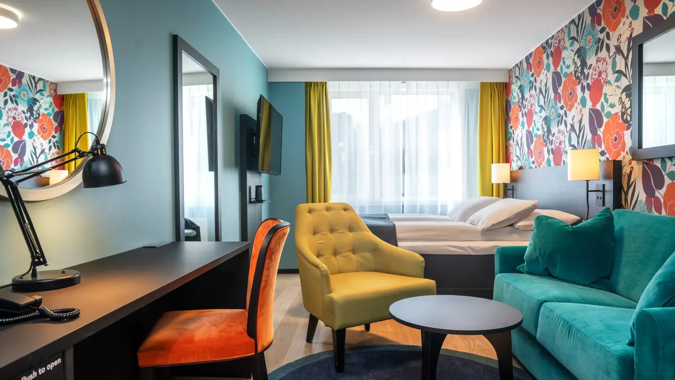 Blomstertapetvegg, turkis sofa, gul stol, salongbord, skrivebord, oransje stol, gulvteppe, parkettgulv, dobbeltseng, vindu, veggspeil, TV, gule gardiner