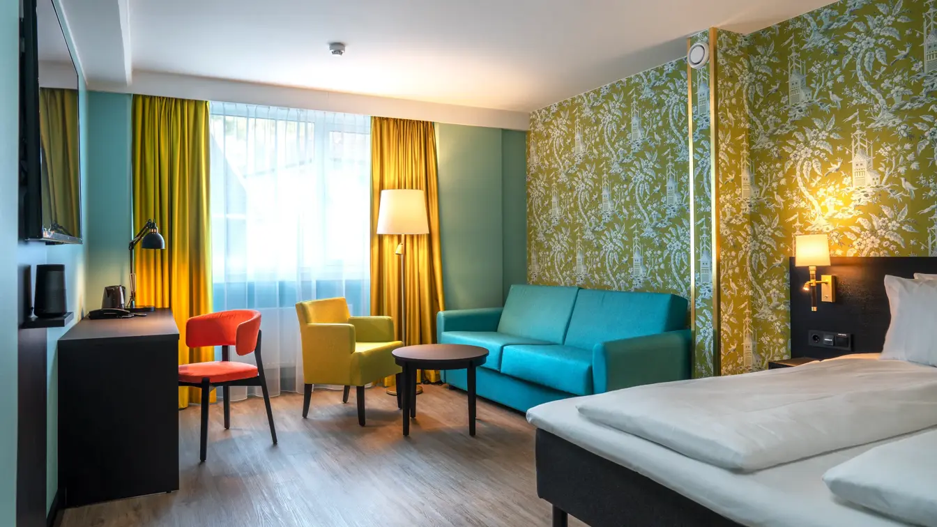Sittegruppe med turkis sofa, sort rundt bord og gul stol. Fargerik mønstret tapet, turkis vegg og gule gardiner. Skrivebord med oransje stol, TV og dobbeltseng i familie rom på Thon Hotel Maritim.