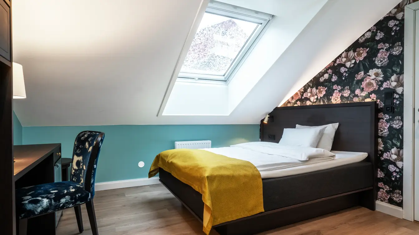 Skråtak med vindu. Mørk tapet med store lyserosa roser. Queen size seng med gult sengeteppe. Mørkt skrivebord med mønstrete stol. I standard rom queen på Thon Hotel Høyers i Skien.