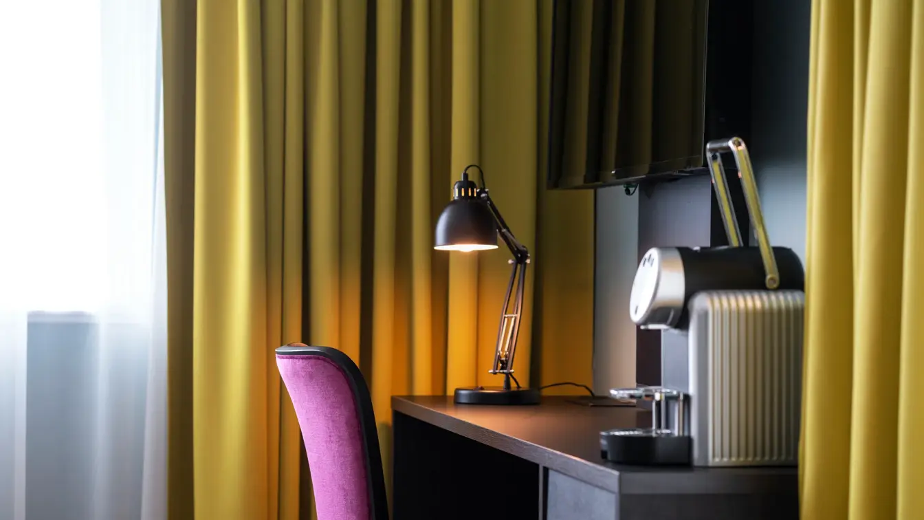 Tv, skrivebord, rosa stol, kaffemaskin, gule gardiner, vindu