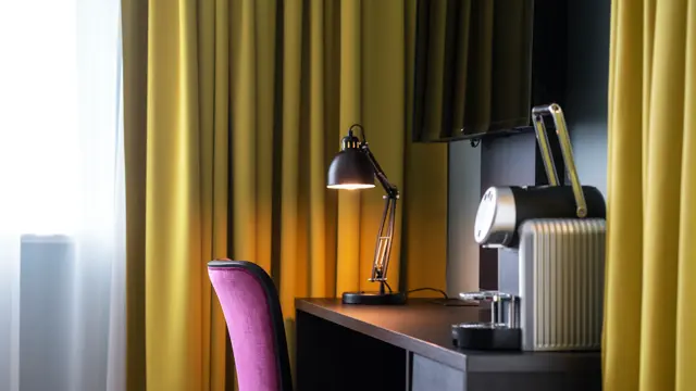 Tv, skrivebord, rosa stol, kaffemaskin, gule gardiner, vindu