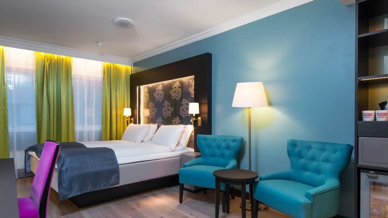 Dobbeltseng,blå lenestoler, gule gardiner og skrivepult med lilla stoler på Thon hotel Terminus i Oslo sentrum nær Jernbanetorget