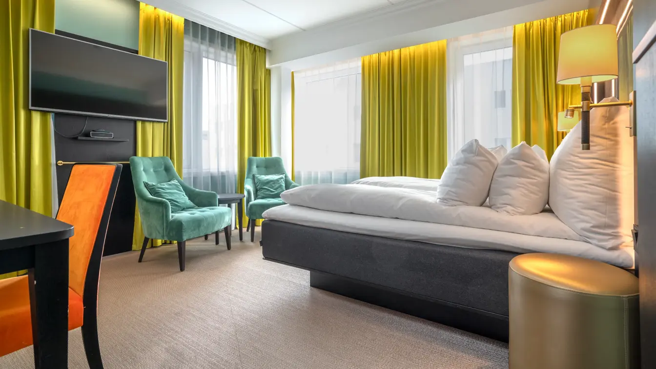 Superiorrom med dobbeltseng, gule gardiner, grønne lenestoler, vegghengt TV.