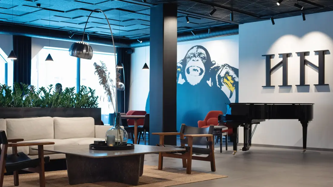Hvit sofa og mørke stoler rundt et lavt sort bord. Hvit vegg med en stort malt ape på.  I lobbyen på Thon Hotel Norge i Kristiansand.