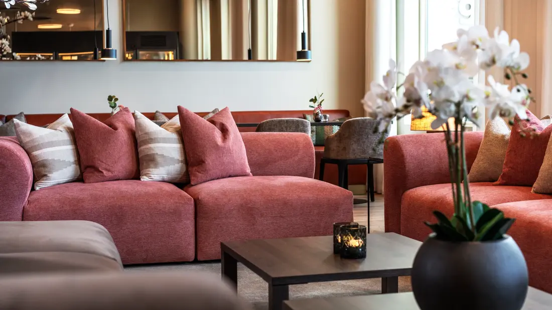 Lyserød sofagruppe med puter og lave salongbord i lobbyområde.