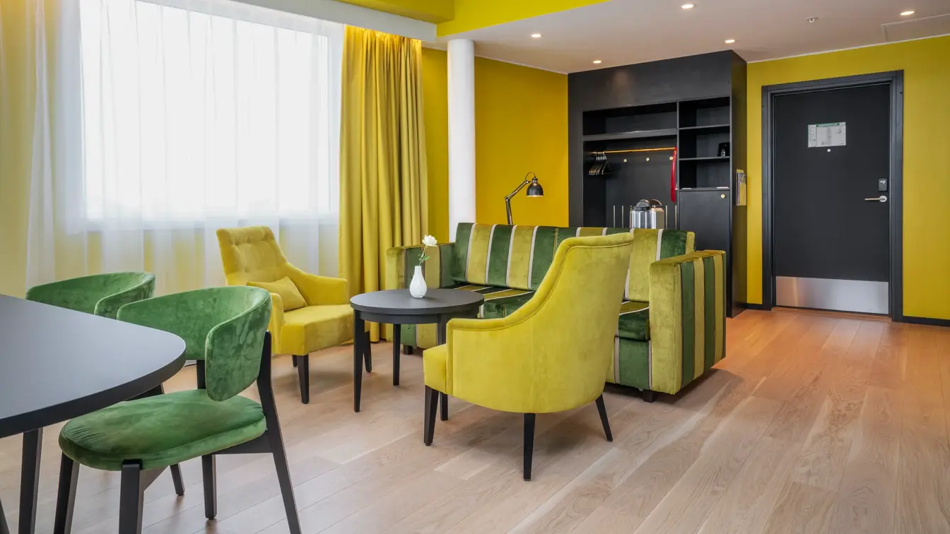 Sittegruppe med gul og grønn stripete sofa, rundt bord, gule stoler, spisegruppe med mørkt rundt bord og grønne stoler, gule vegger og gule gardiner i suite på Thon Hotel Kirkenes