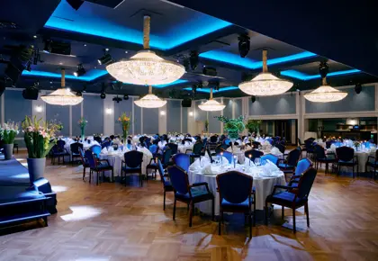 Selskapslokale dekket opp med runde bord dekket med hvite duker og servise. Blå stoler, parkett gulv og store lysekroner i taket med kald, dempet belysning. En opphøyd svart scene til venstre med blomster på hver side.