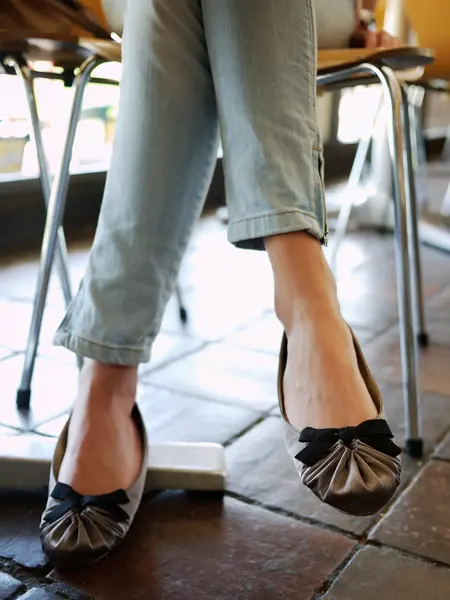 Nærbilde av dameben med jeans og ballerinasko med svart sløyfe, sittende på en stol. 