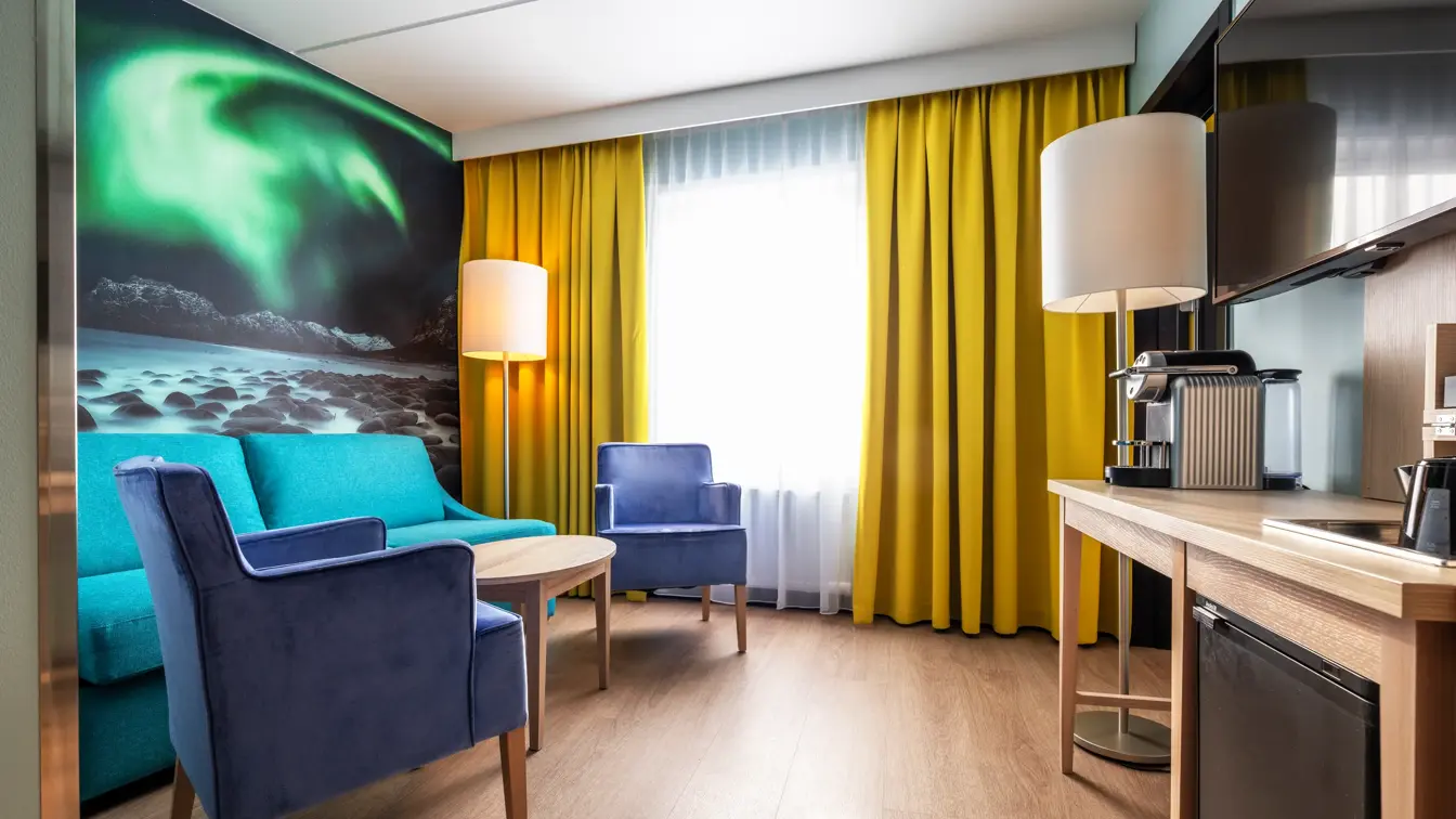 Junior suite med sittegruppe, stort bilde av nordlys på veggen, gule gardiner, bord med Nespresso maskin og vegghengt TV skjerm over.