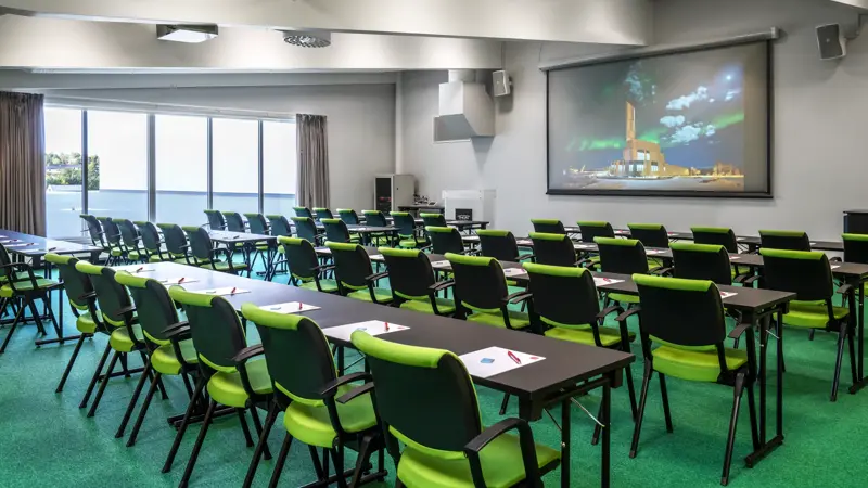 Skrivebordpulter, hvit vegg med projektorlerret, vindu med grå gardiner, vindu fra gulv til tak, grønt teppegulv, grønne stoler.  